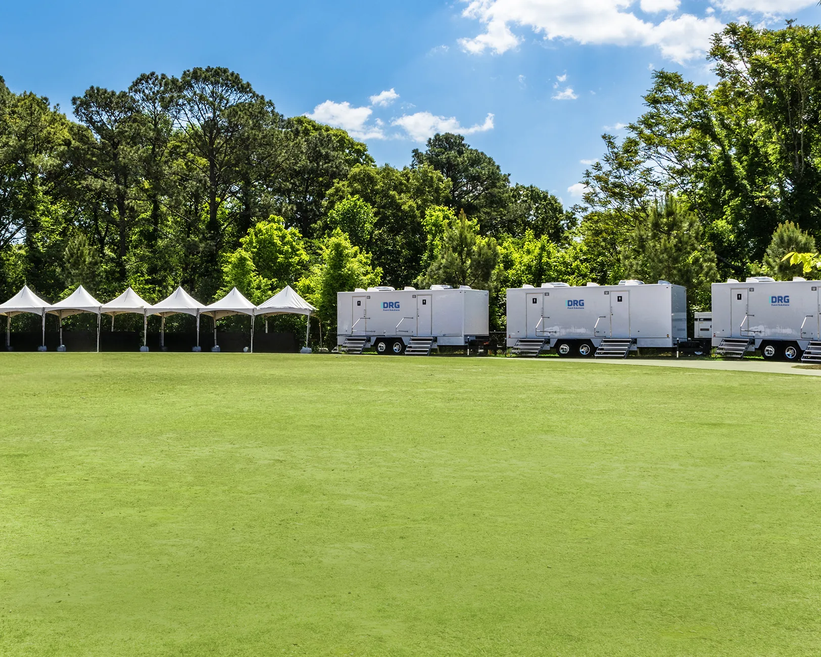White DRG restroom trailers and a row of white event tents along the edge of a green field.
