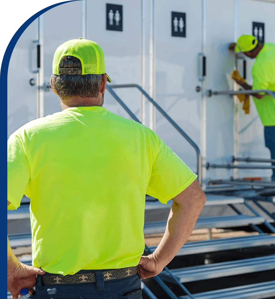 DRG crew member in a high-visibility shirt and cap watching a colleague service a restroom trailer.