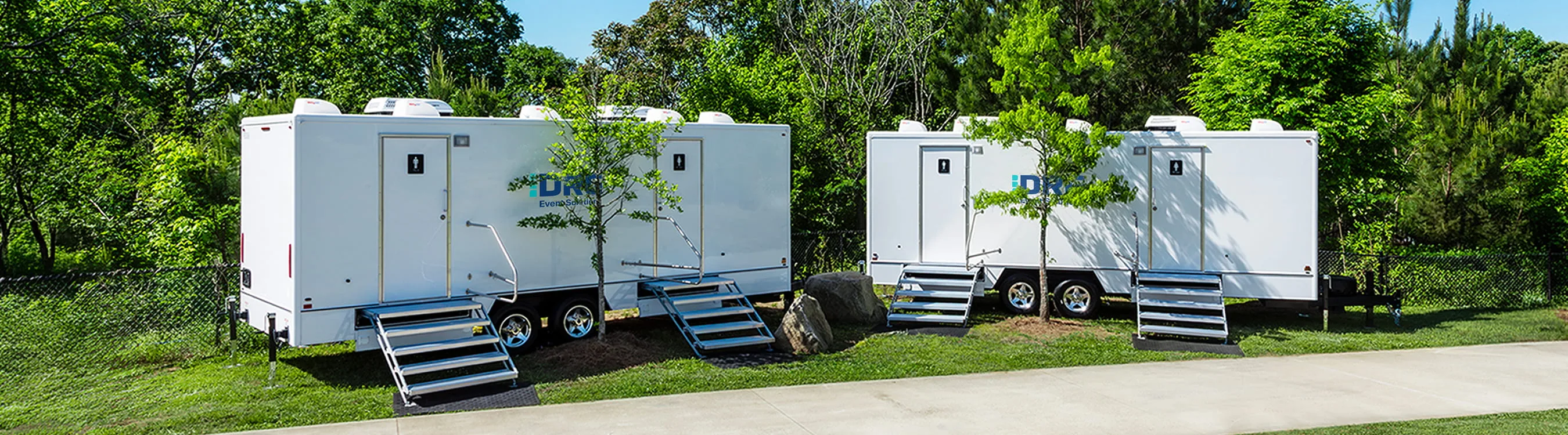 Two white DRG restroom trailers with aluminum stairs parked on a grassy lot beside trees.