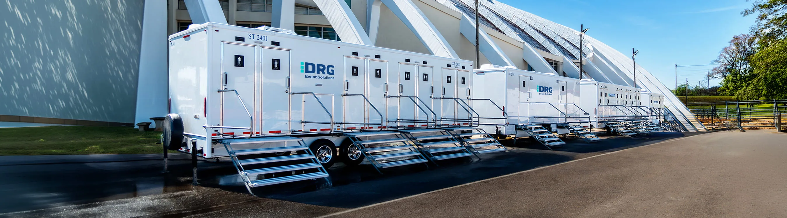 A long row of white DRG shower trailers with stairs parked outside a stadium.
