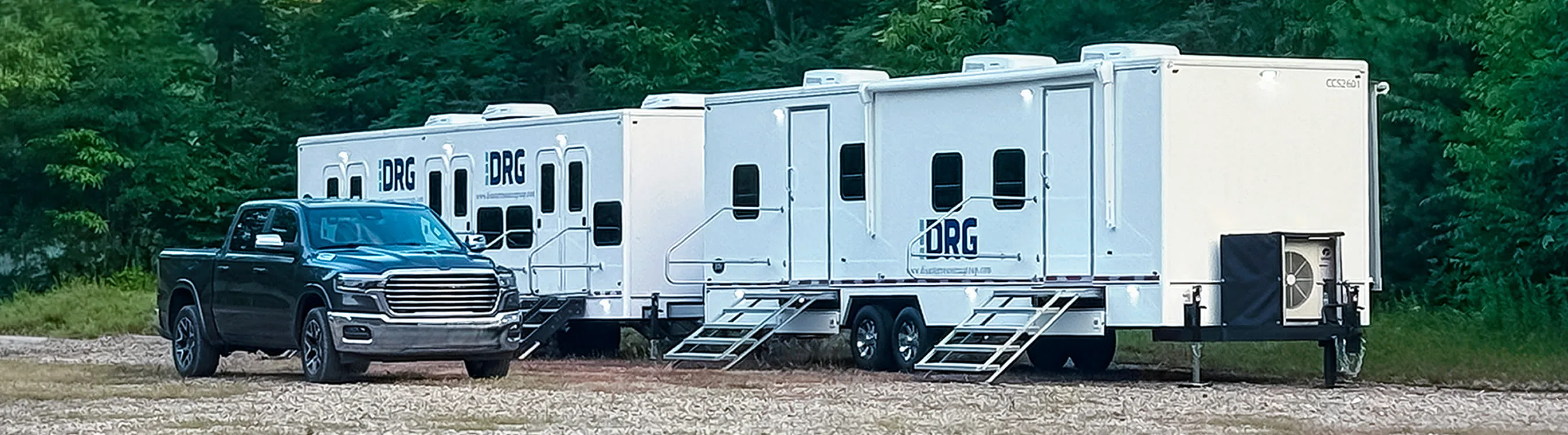 Large white DRG specialty trailers with stairs parked on gravel next to a pickup truck.