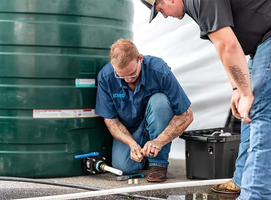 Two DRG technicians connecting hoses and fittings at the base of a large green water tank.