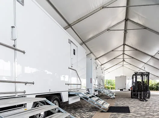 Restroom trailers lined up inside a large white event tent with a forklift in the background.