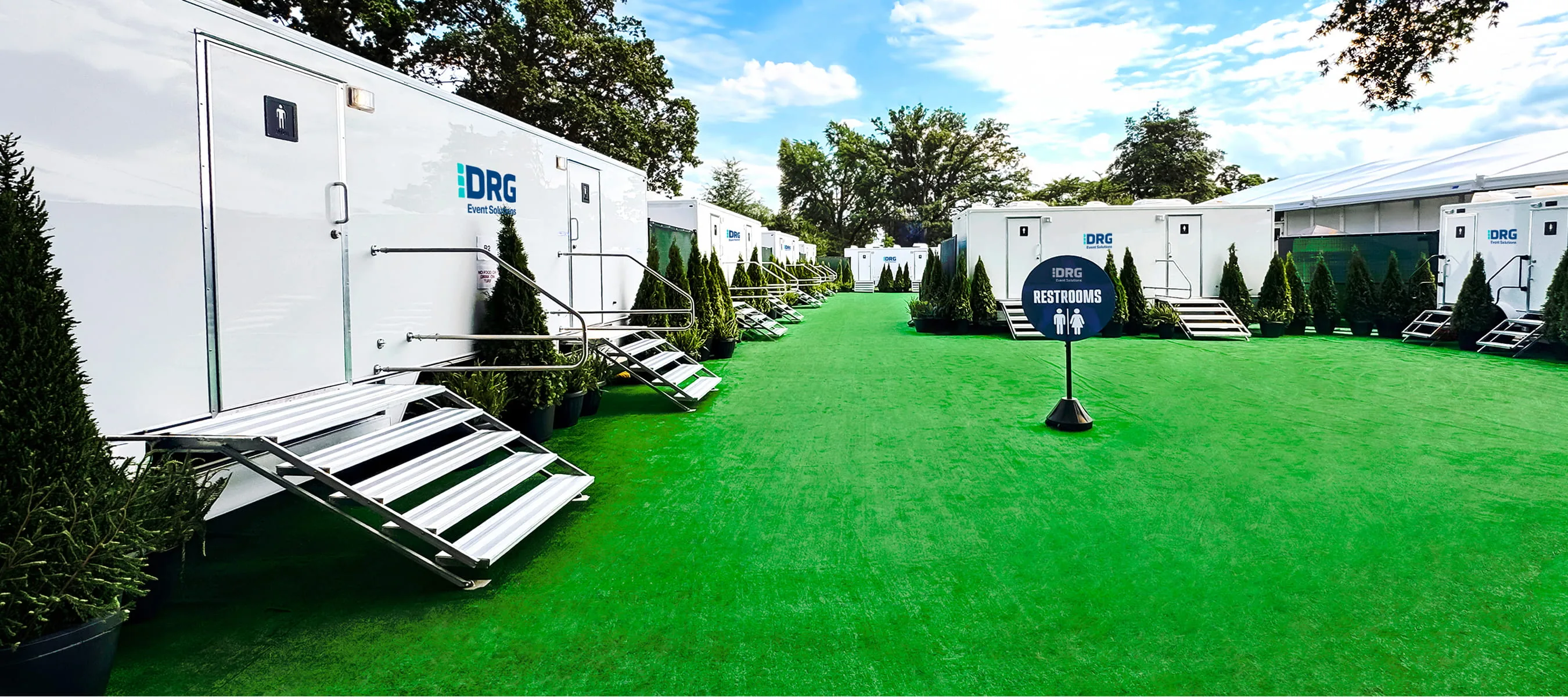 Long rows of white DRG Event Solutions restroom trailers on artificial turf with a restrooms sign.