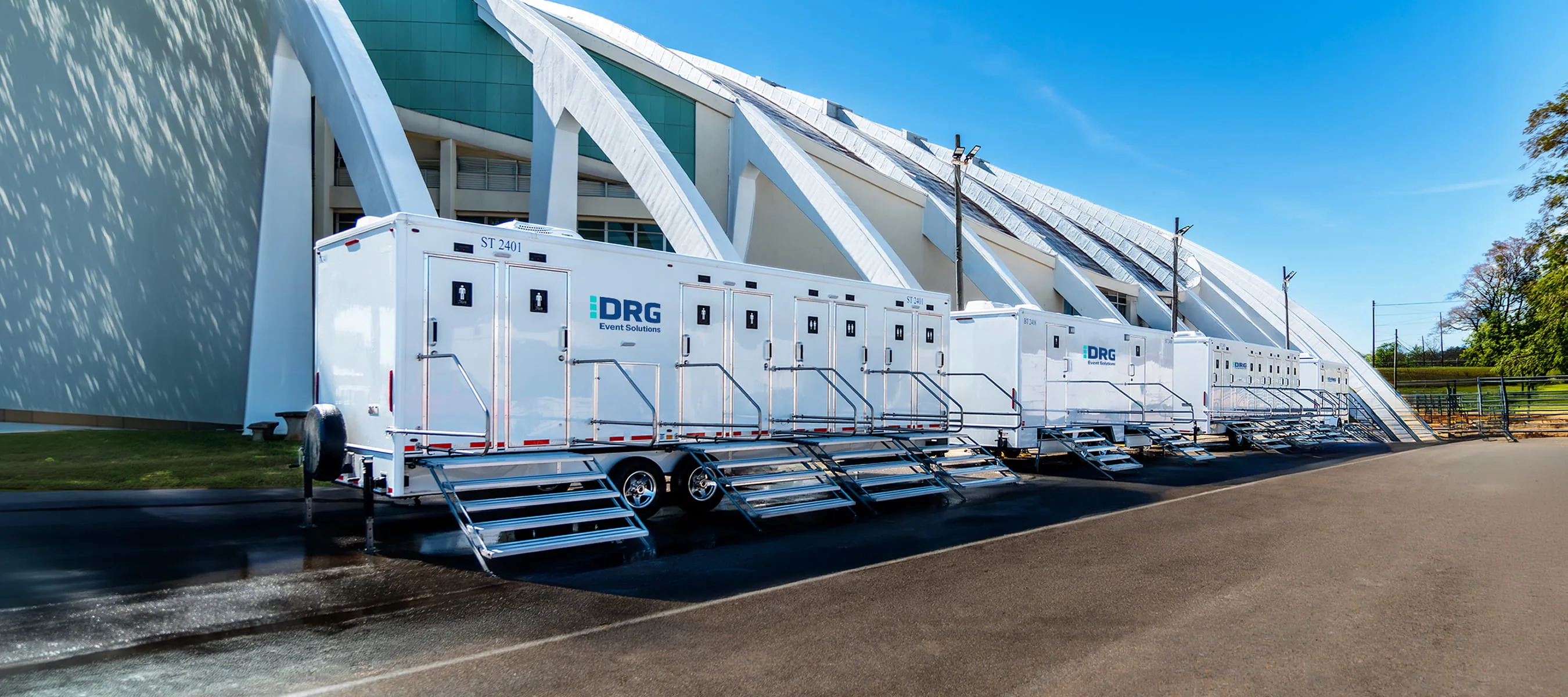 A line of white restroom trailers parked along the curved facade of a stadium.