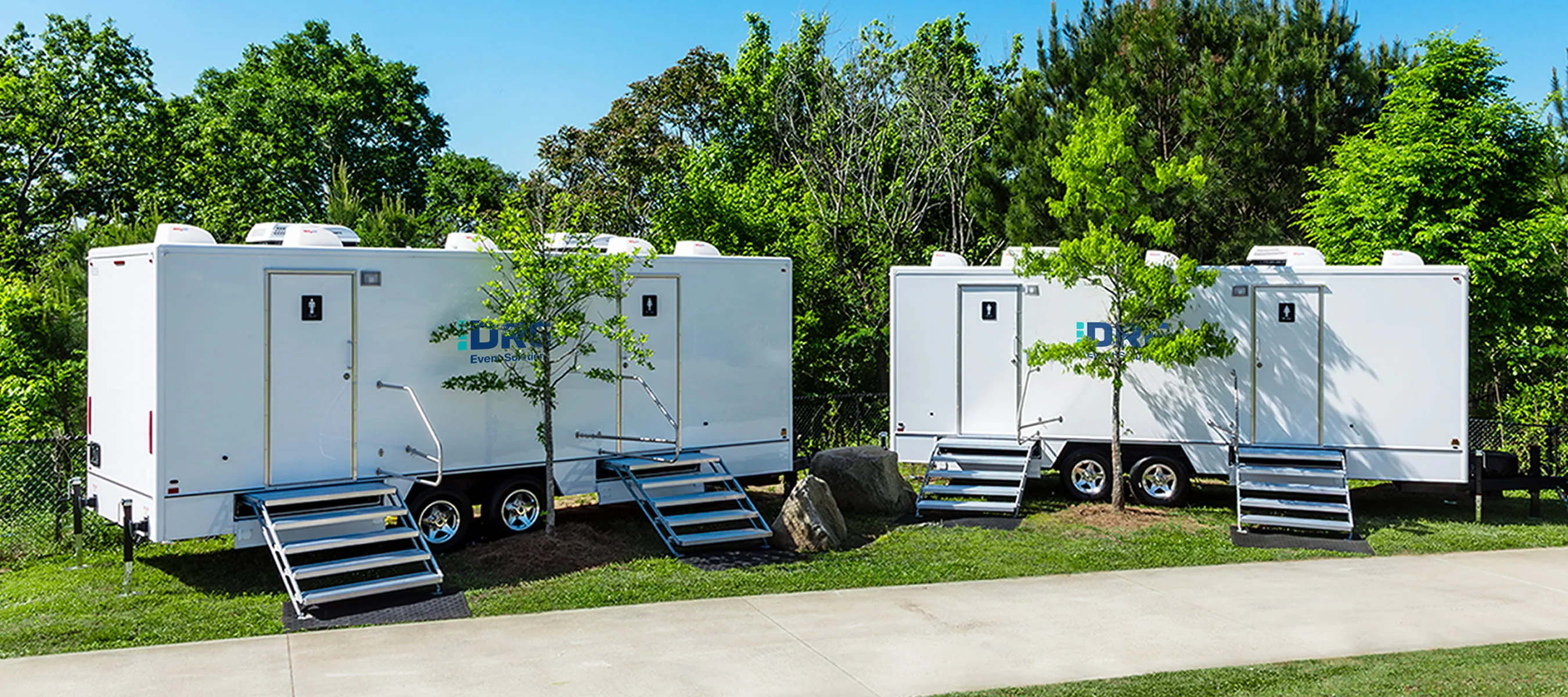 DRG Event Solutions restroom trailers with stairs set up on grass under tall trees.