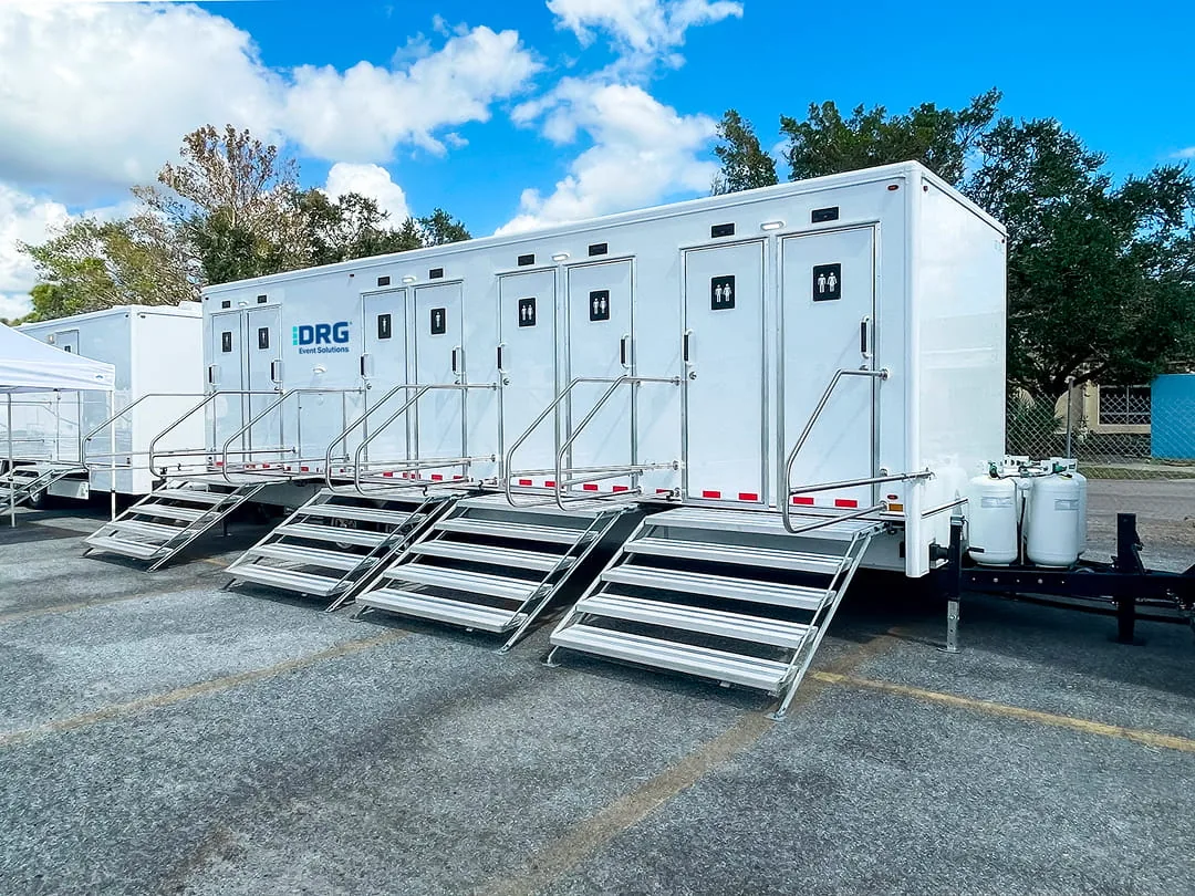 Row of white DRG Event Solutions restroom trailers with aluminum stairs parked on a paved lot.