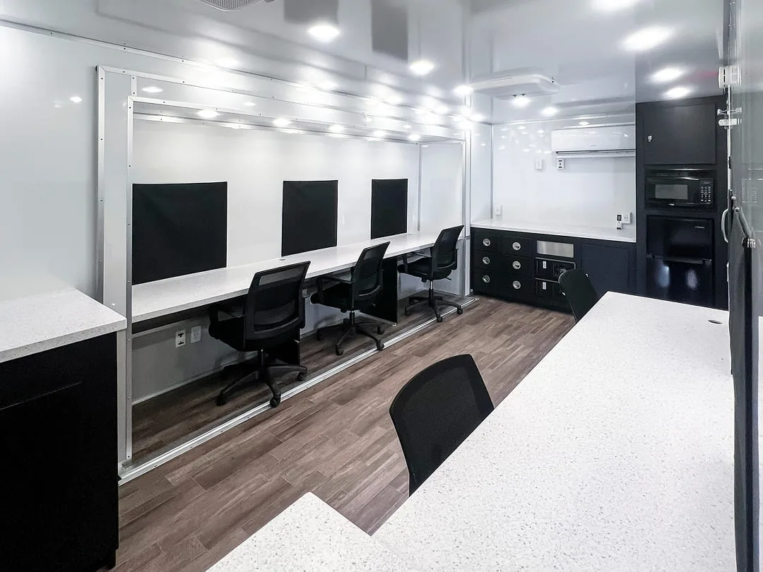 Command trailer interior with a long white work counter, office chairs, and wall-mounted monitors.