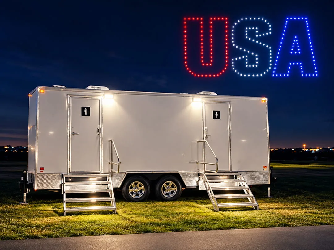 Restroom trailer with lit stairs parked on a field at night beneath a red, white, and blue USA light display.