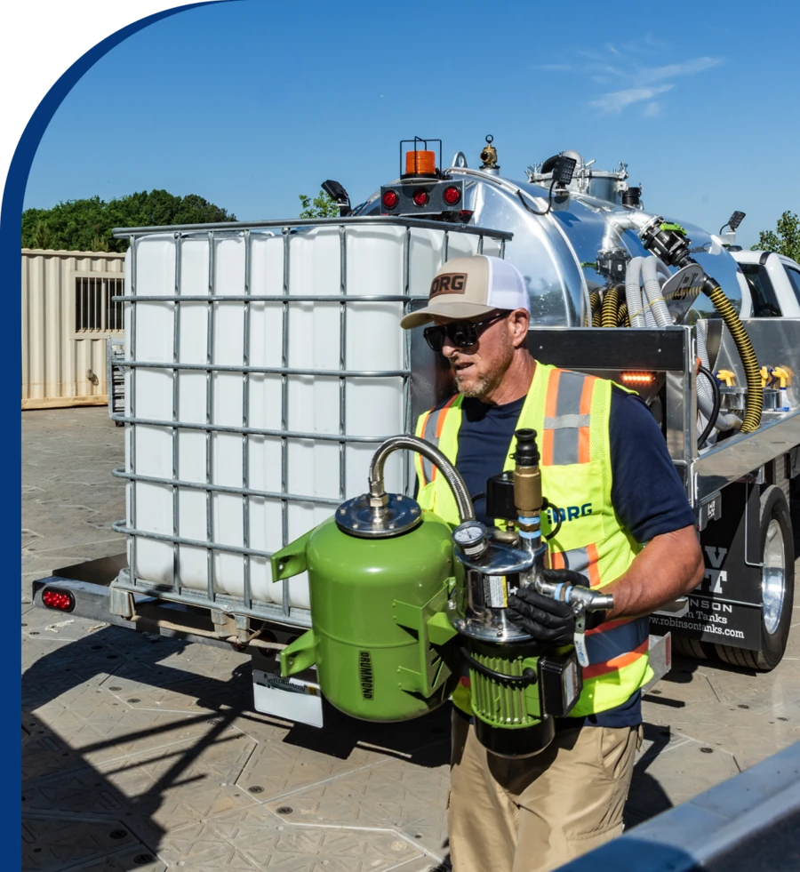 DRG service technician in a high-visibility vest carrying pump equipment past a water tank and service truck.
