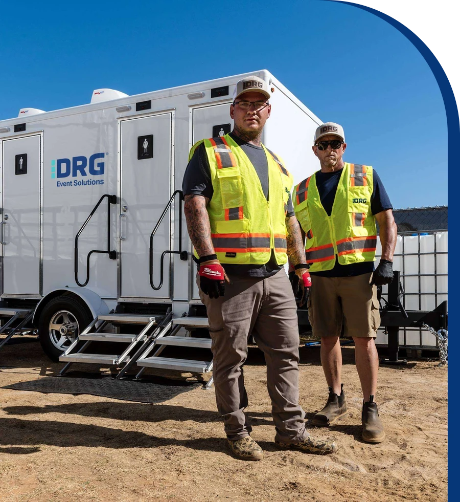 Two DRG crew members in safety vests and caps standing in front of a restroom trailer.